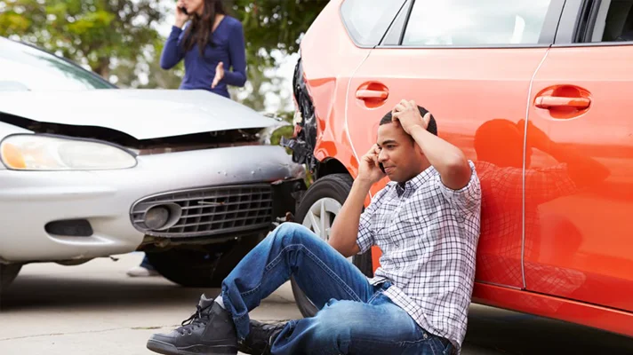 person sitting on the ground on cellphone next to a car collision