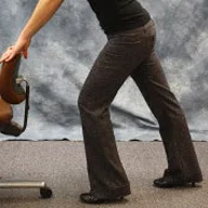 person standing slightly bent with hands on the back of a chair, left leg forward and slightly bent, right leg straight